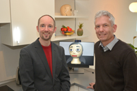 Dr. Thomas Hermann (left) und PD Dr. Sven Wachsmuth (right) are heading the project together with Prof. Dr. Britta Wrede. The avatar Flobi (center) provides support in the kitchen to the apartment’s guests. Photo: CITEC/Bielefeld University