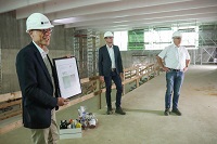Bild: Kanzler Dr. Stephan Becker mit zwei Mitarbeitern der Firma Terhalle Holzbau GmbH unter der hölzernen Decke des neuen Hörsaalgebäudes. Mit einer Grundfläche von über 600 qm und einer Deckenhöhe von bis zu knapp 10 Metern bietet der große Hörsaal zukünftig Platz für Vorlesungen mit bis zu 650 Zuhörer*innen.  